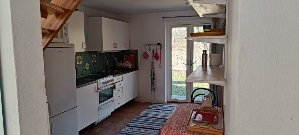 bild på ett avlångt kök med glasad backdörr. ett litet vord och hyllor. kyl och frys staplade på varandra. 