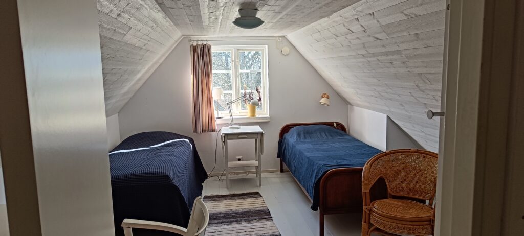 två sängar och ett litet sängbord. två fotöljer och en matta. snedtag och fönster ut mot baksidan
